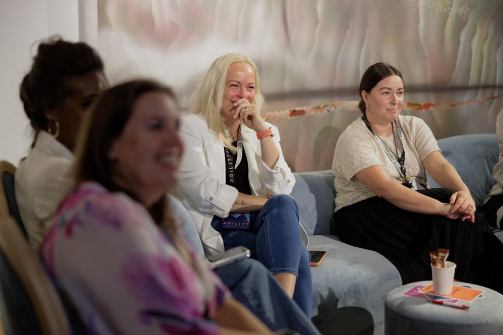 Three women sit on a blue sofa in a bright room, with a blonde woman in a white blazer laughing while others listen during a professional event.