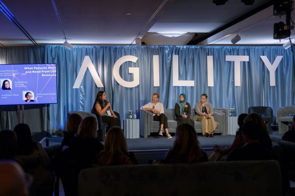 A panel discussion on a stage with the word AGILITY in large letters on a blue curtain backdrop, featuring four speakers seated on a grey sofa.