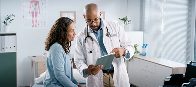 A doctor shows a tablet to a seated patient in a medical exam room. The doctor wears a white coat and stethoscope; the patient listens attentively.
